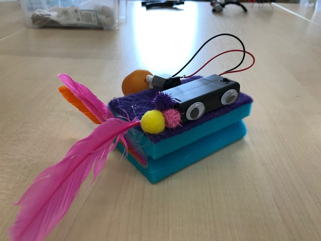 Kleines selbstgebautes elektronisches Gerät mit bunten Federn, Pompons und zwei Kabeln, Ergebnis eines Coding & Making Workshops
