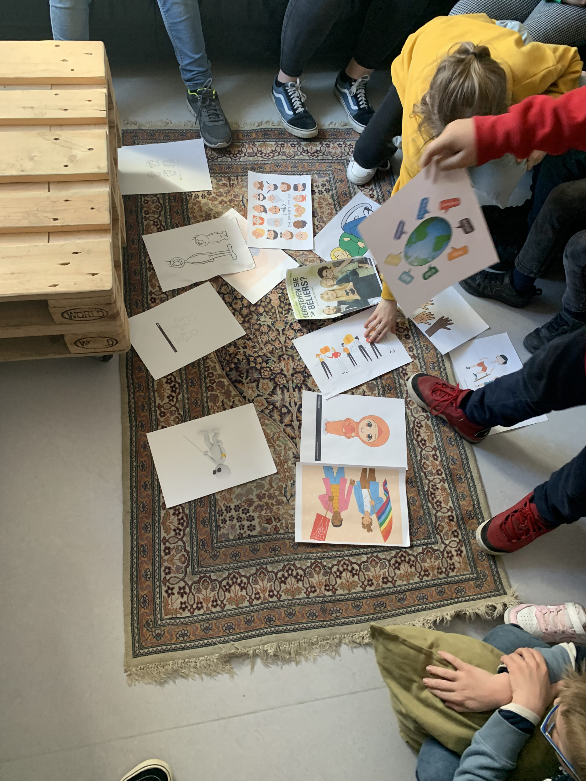 Verschiedene Bilder, die Diskriminierungskategorien darstellen, liegen auf einem Boden. Drum herum stehen Menschen und schauen sich die Bilder an.