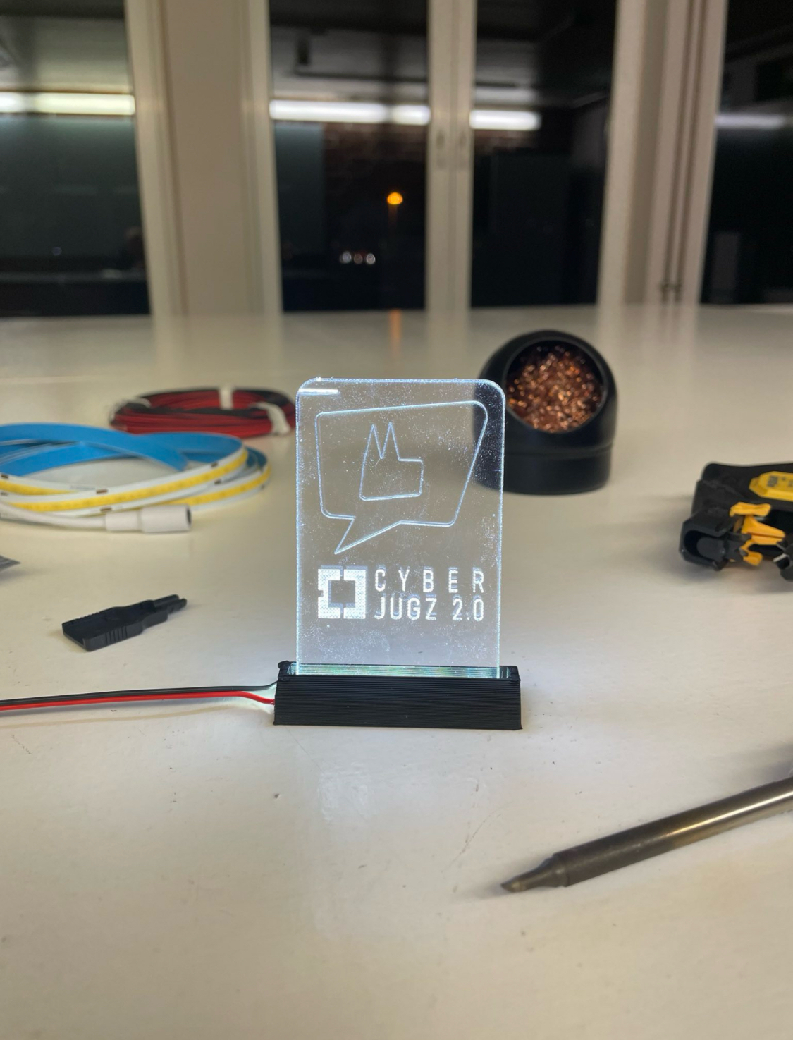 Leuchtendes Acrylschild mit Aufschrift 'CYBER JUGZ 2.0' auf einem Tisch mit Werkzeugen und Kabeln im Hintergrund