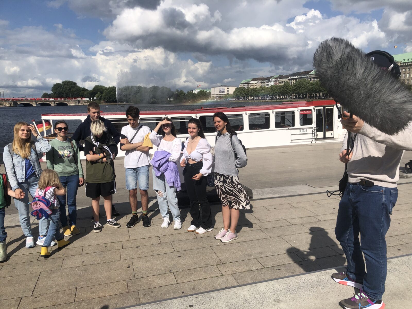 Eine Gruppe von Jugendlichen steht zusammen an einem Hafen, ganz vorne steht einer und hält ein großes Mikrofon nach oben.