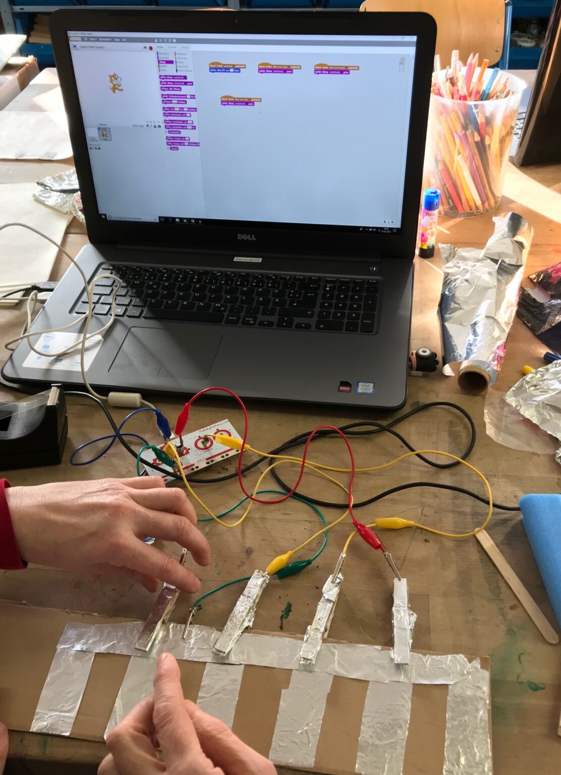 Ein Laptop steht auf einem Tisch, darauf ist das Scratch Programm geöffnet. Ein MakeyMakey ist angeschlossen und wird mit einer selbst gebastelten Tastatur verbunden.