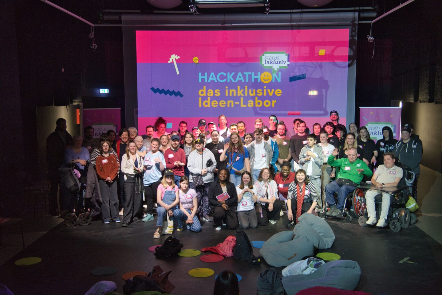Teilnehmende vom Hackathon stehen gemeinsam auf der Bühne und jubeln. Dahinter ist eine Leinwand auf der steht "Hackathon das inklusive Ideen-Labor"