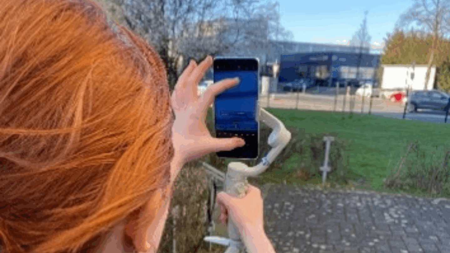 Eine Jugendliche hält ein Gimbal mit einem Smartphone in der Hand und filmt draußen eine Szene.