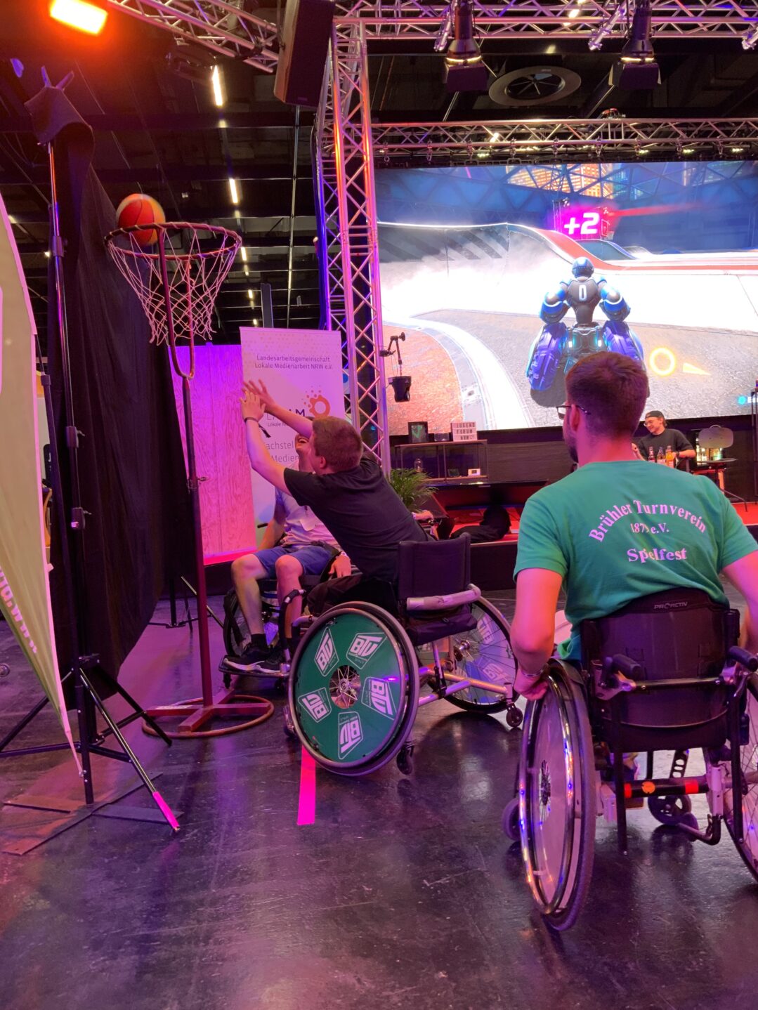 Eindrücke vom Jugendforum NRW 2025: Menschen spielen vor der Bühne echten Rollstuhl-Basketball und werfen gerade einen Korb. Auf der Bühne wird an der Konsole digital Rollstuhl-Basketball gespielt, was man auf einem großen Screen sieht.