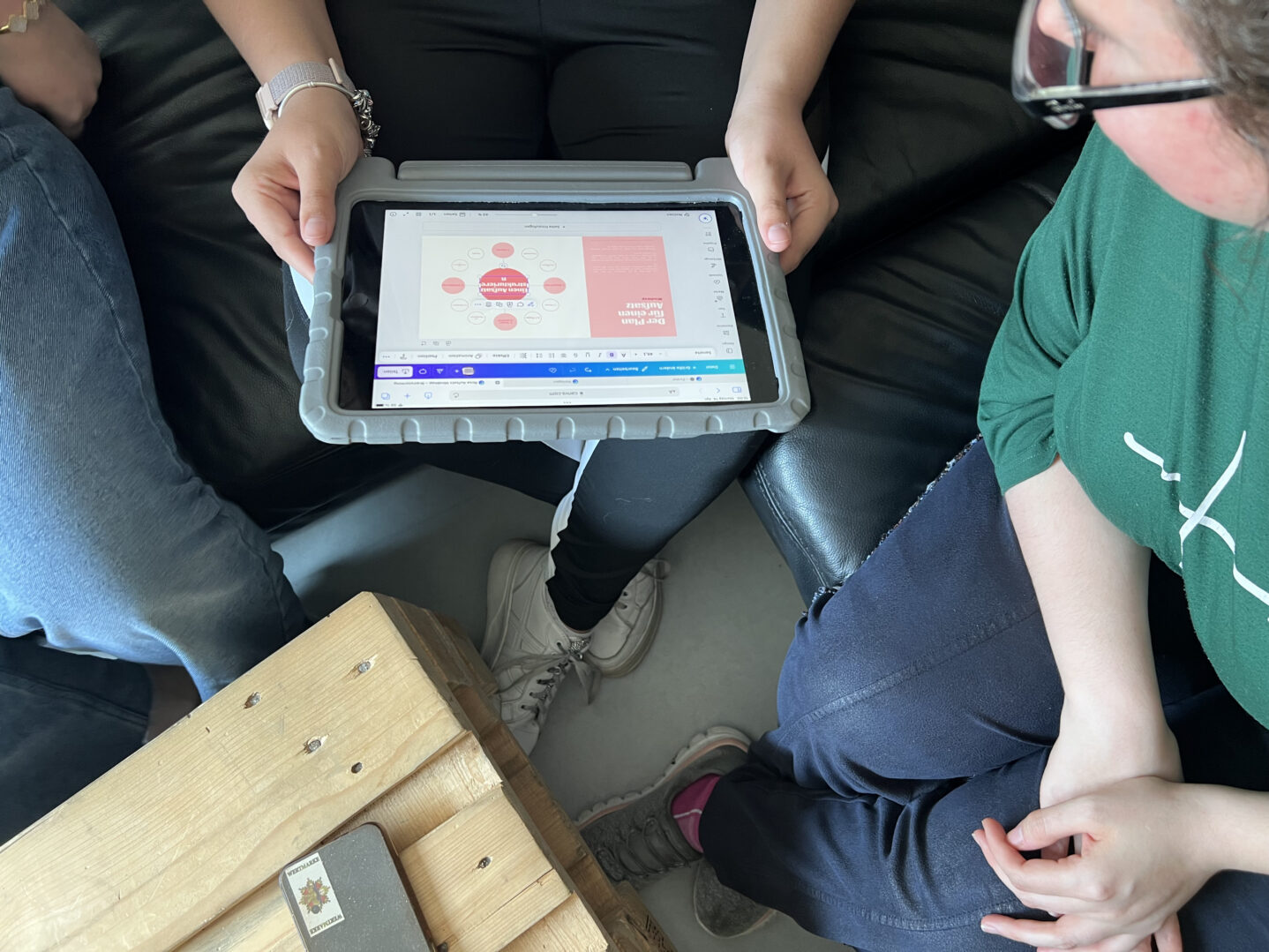 Eine Person hält ein Tablet mit einer grafischen Darstellung einer Mindmap in der Hand. Auf dem Tablet sind mehrere rote und rosa Kreise mit Text zu sehen. Zwei weitere Personen sitzen auf einem Sofa und schauen auf das Tablet.