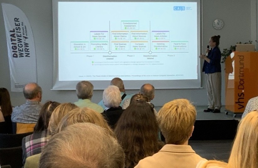 Der Blick über die Köpfe des Publikums auf einen Vortrag auf der Bühne mit einer Grafik auf dem Beamerbild an der Leinwand.