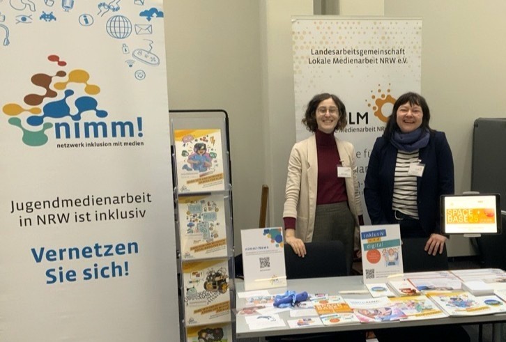 Der Stand der LAG LM mit zwei Team-Mitgliedern auf dem Fachtag mit Magazinen und Aufstellern. Das Roll-Up im Hintergrund sagt "nimm! Netzwerk Inklusion mit Medien. Jugendmedienarbeit in NRW ist inklusiv. Vernetzen Sie sich!"
