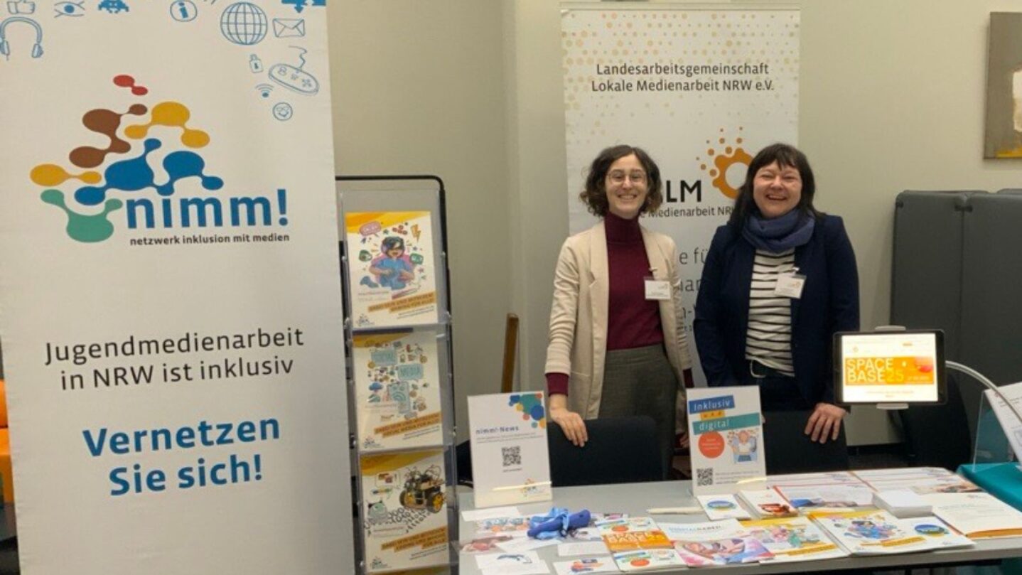Der Stand der LAG LM mit zwei Team-Mitgliedern auf dem Fachtag mit Magazinen und Aufstellern. Das Roll-Up im Hintergrund sagt "nimm! Netzwerk Inklusion mit Medien. Jugendmedienarbeit in NRW ist inklusiv. Vernetzen Sie sich!"
