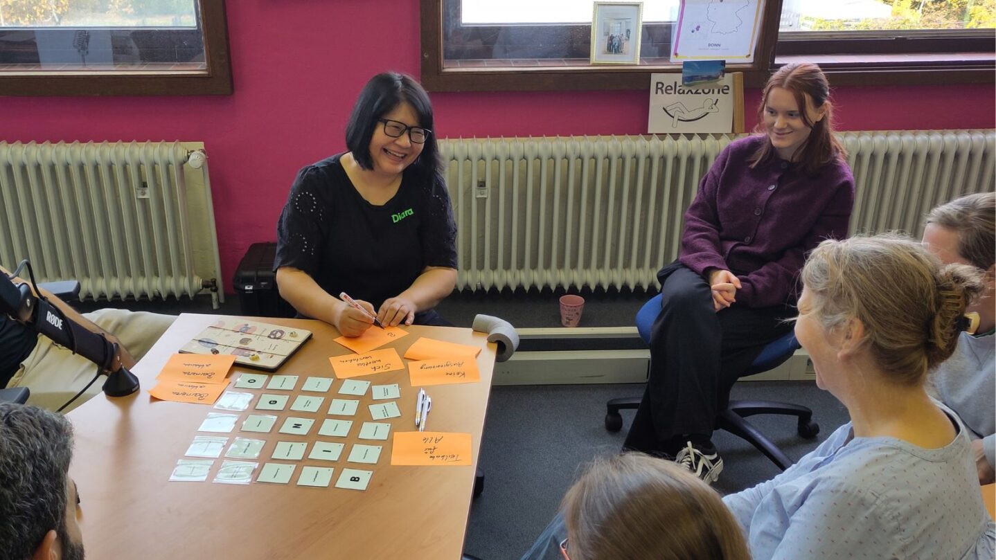 Fachkräfte sitzen in einer Stuhlgruppe und lachen, während sie auf Zetteln Notizen machen.