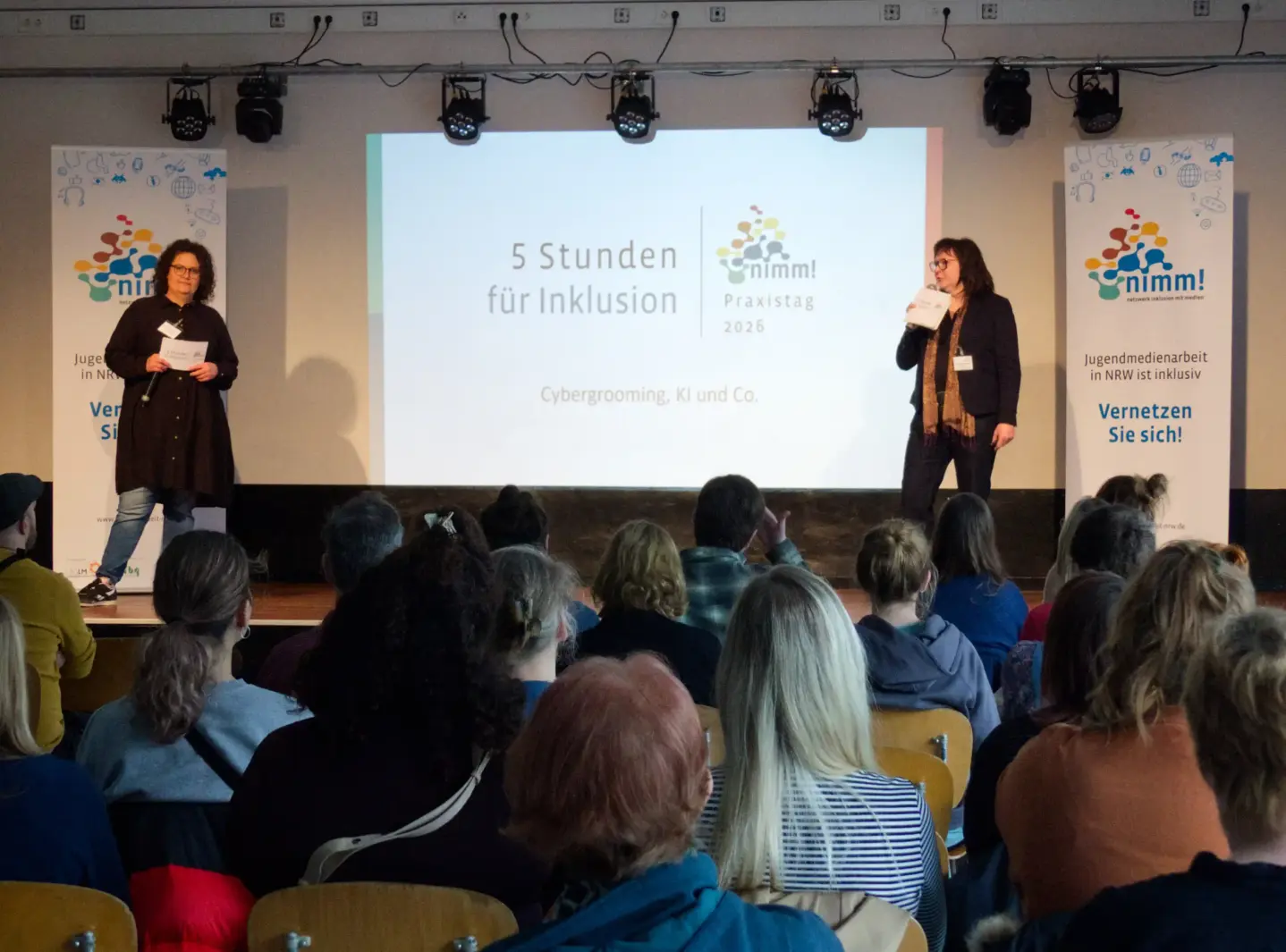 Zwei Personen stehen auf einer Bühne und präsentieren etwas. Vor der Bühne sitzt ein Publikum.