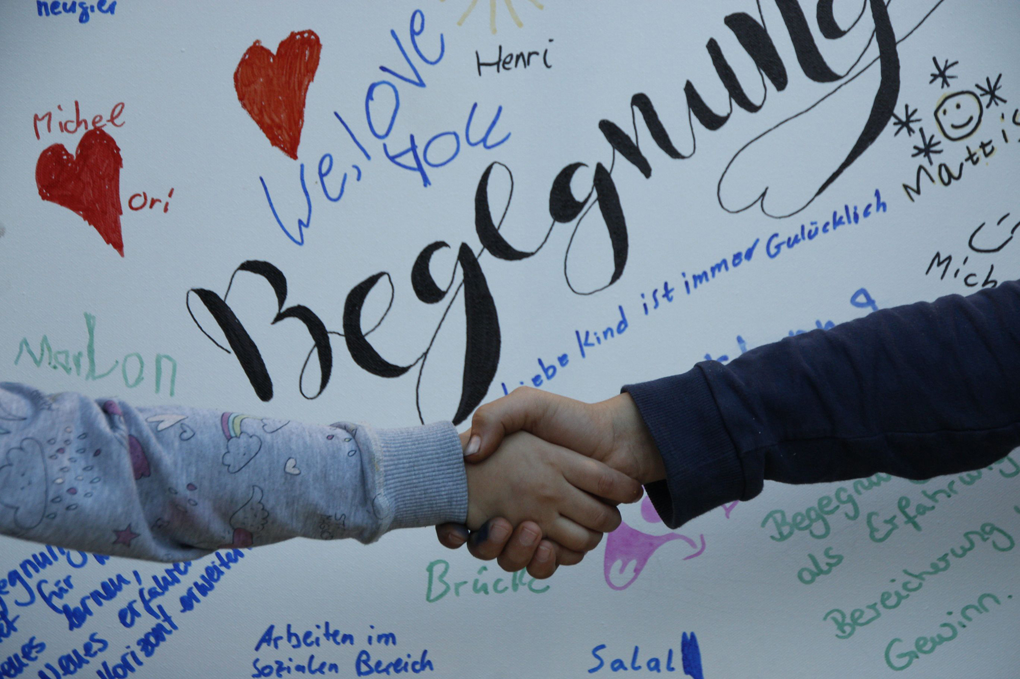 zwei Hände, die sich zur Begrüßung greifen. Dahinter eine Wand auf der groß Begegnung steht.