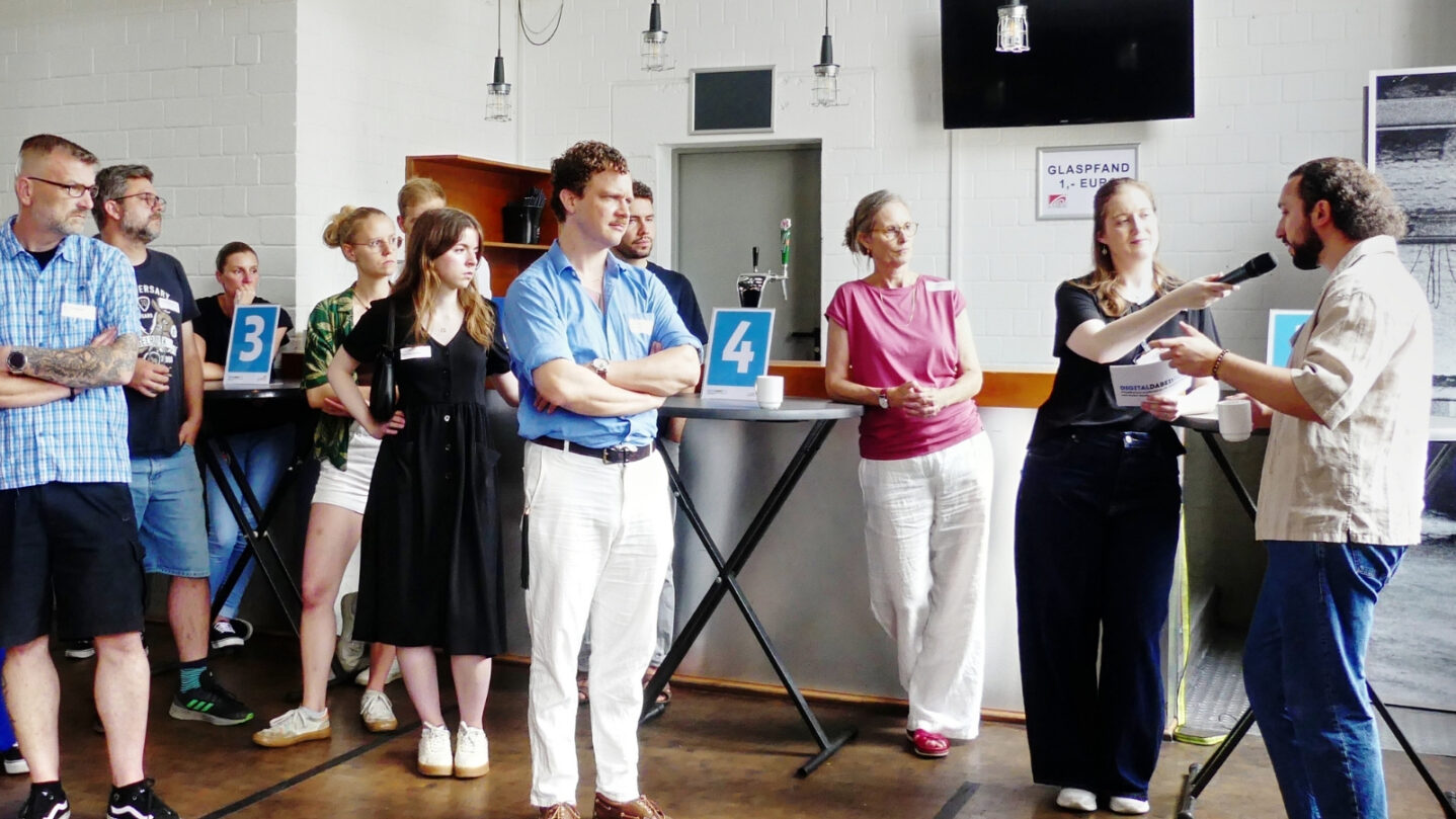 Gruppe von Menschen steht in einem Raum, eine Frau hält ein Mikrofon zu einem Mann und interviewt ihn