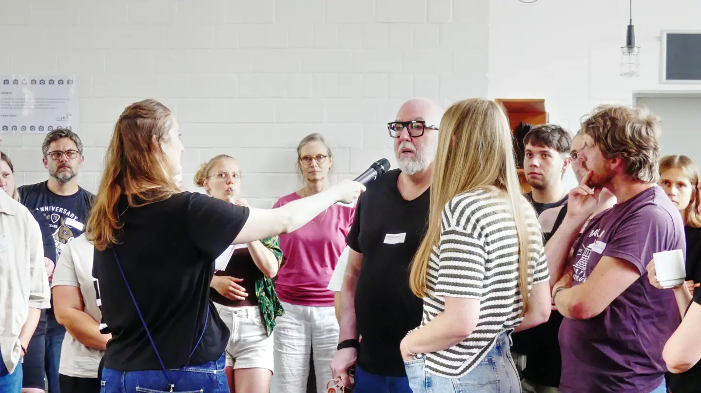 Gruppe von Menschen in einem hellen Raum, eine Frau hält ein Mikrofon zu einem älteren Mann mit Brille und Bart, andere hören zu und schauen interessiert.